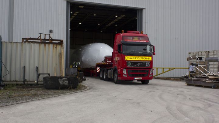 A red truck exits a warehouse, carrying a large, covered object, with workers and equipment nearby.