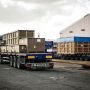 A truck loaded with large crates is parked at a warehouse, with additional trailers visible in the background.