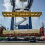 A crane lifts a container at a shipping yard under a bright sky, surrounded by cargo and port equipment.