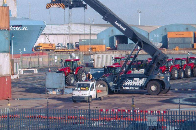 A crane lifts a container at a busy shipping yard with tractors parked nearby and a small vehicle in the foreground.